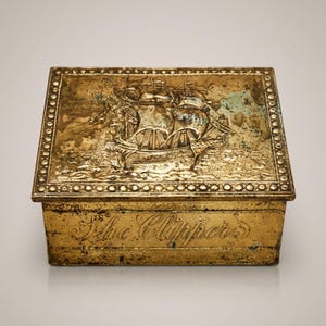 May include: A rectangular brass box with a detailed relief of a sailing ship on the lid. The box has a decorative border and the words "The Clippers" are inscribed on the front. The box has a weathered appearance.