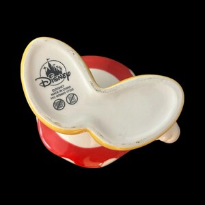 May include: A ceramic Minnie Mouse-themed mug, featuring a white interior and a red and white striped exterior. The mug has a yellow handle and the Disney logo is visible on the bottom. The mug is made in China.