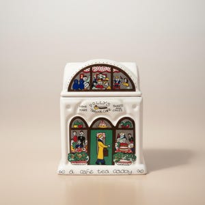 May include: A white ceramic tea caddy shaped like a cafe building. The front features colorful illustrations of people, cakes, and a green door. Text reads: Polly's Home Made Teapot Cafe Sweets and Cakes, and at the bottom, a cafe tea caddy.