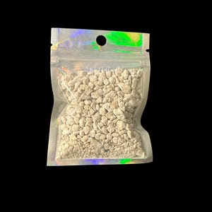 May include: A clear, iridescent zip-top bag filled with small, light-colored pebbles. The bag has a small hole near the top and is set against a black background. The pebbles appear to be a natural material.