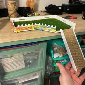 May include: A white and green cat toy with a grass-like surface and a picket fence design. A roll of brown rope is held in a hand. The toy is on a wooden surface with craft supplies.