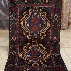 Puede incluir: Una alfombra rectangular anudada a mano con un campo central azul marino con patrones geométricos en marrón, rojo y beige. La alfombra tiene un borde oscuro con motivos repetidos y flecos en el borde superior. La alfombra se exhibe sobre un suelo claro.