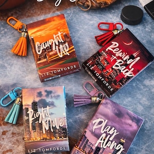 May include: Four rectangular book-themed bag charms featuring cityscapes and titles by Liz Tomforde. Each charm includes a colorful tassel and a matching lobster clasp. The titles are Caught Up, Rewind it Back, The Right Move, and Play Along.