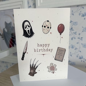 May include: A white greeting card with the text happy birthday in the centre, surrounded by illustrations of a ghost mask, a hockey mask, a red balloon, a bloody knife, a clawed glove, a book titled Handbook for the Recently Deceased, and a puzzle piece.