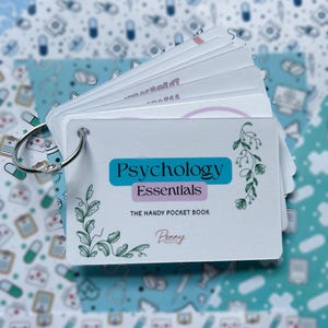 May include: A set of white flashcards bound by a silver metal ring. The front card features the text Psychology Essentials, The Handy Pocket Book, and Penny Paper Co. in black and blue, decorated with green botanical illustrations on the sides.