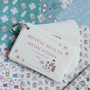 Könnte beinhalten: Ein weißes Karteikartenset, das mit einem silbernen Metallring zusammengehalten wird. Die vordere Karte zeigt den Text MENTAL HEALTH MEDICATIONS, The handy pocket book und Penny, verziert mit kleinen Blumenmotiven und Abbildungen von Pillendosen.