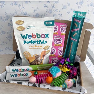 May include: A cardboard gift box containing various cat treats and toys. Items include a white bag of Webbox Pocketfuls with cheese, a pink package of Bonkers Purr Pops, a teal stick of Big Pack treats, and two Webbox liver sausage and chicken sticks.