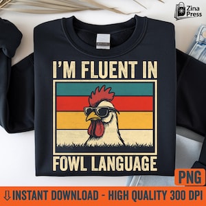 May include: Black crewneck sweatshirt with the text "I'M FLUENT IN FOWL LANGUAGE". The graphic features a rooster wearing sunglasses, set against a retro color block of teal, red, yellow, and orange.