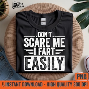 Könnte beinhalten: Schwarzes T-Shirt mit dem weißen Text "DON'T SCARE ME I FART EASILY". Das Shirt liegt auf einer gewebten Matte, mit Jeans und einem Turnschuh im Hintergrund. Das Bild enthält auch eine "PNG"-Grafik.