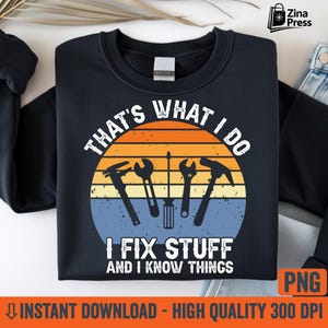 May include: Navy blue sweatshirt with a circular graphic featuring tools and the text "THAT'S WHAT I DO, I FIX STUFF AND I KNOW THINGS." The graphic has a sunset design with orange, yellow, and blue hues. The sweatshirt is a casual garment.