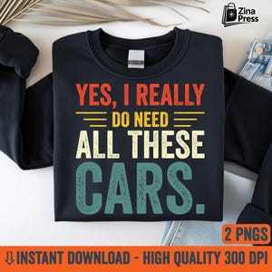 May include: Black sweatshirt with the text "YES, I REALLY DO NEED ALL THESE CARS." in red, yellow, and teal lettering. The sweatshirt is displayed on a white surface with a pair of jeans visible in the background. The image also includes the text "2 PNGS" and "INSTANT DOWNLOAD - HIGH QUALITY 300 DPI."