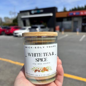 May include: A glass jar candle with a wooden lid. The label reads "WHITE TEA & SPICE soy wax candle." The candle is hand-poured and contains 198 grams of wax. The label features floral and spice illustrations.