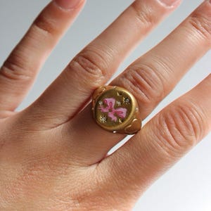 May include: A gold ring featuring a pink bow design, small gemstones, and heart and star details. The ring is worn on a finger with a French manicure. The ring's design is set on a gold base.
