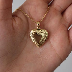 Puede incluir: Un collar con un colgante en forma de corazón dorado. El medallón tiene un diseño ornamentado y cuelga de una delicada cadena. El collar se sostiene en una mano abierta.