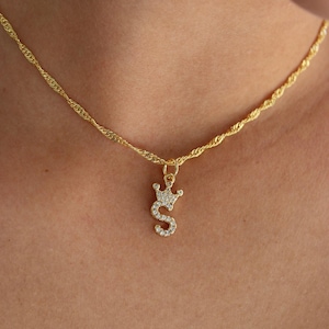 May include: A gold-colored necklace with a pendant shaped like the letter "S" topped with a crown. The pendant and crown are encrusted with small, clear gemstones. The chain has a twisted rope design.