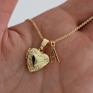 Puede incluir: Collar con colgante de medallón en forma de corazón dorado y una inicial. El medallón tiene un diseño ornamentado y se abre para revelar un espacio para una foto o un pequeño recuerdo. La cadena del collar es delicada.