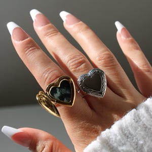 Könnte beinhalten: Zwei herzförmige Medaillonringe, einer goldfarben und einer silberfarben, auf einer Hand mit weiß lackierten Nägeln. Der goldfarbene Ring ist geöffnet und zeigt ein kleines Foto. Der Hintergrund ist ein weiches Grau.