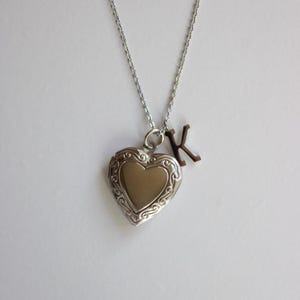 May include: Silver heart-shaped locket necklace with an engraved border and a small letter "K" charm. The locket hangs from a delicate chain, creating a romantic and personalized accessory.