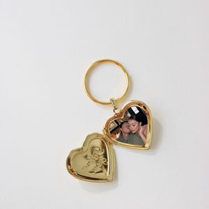 May include: A gold heart-shaped locket with a photo inside, open to reveal the image. The locket is attached to a gold ring. The closed side of the locket has an embossed floral design.
