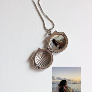 Puede incluir: Collar con colgante de concha plateada y cadena. El relicario está abierto, revelando una pequeña fotografía de una pareja. También es visible una foto separada de la pareja en una playa. Sobre un fondo blanco.