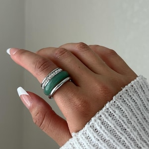 May include: A stack of three rings worn on a finger. The top ring is a thin silver band set with small clear stones. The middle ring is a smooth, rounded green band. The bottom ring is a plain, polished silver band with a rounded profile.
