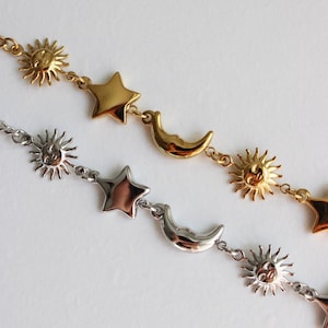 May include: Two celestial-themed necklaces. One is gold-toned, featuring sun, star, and crescent moon charms. The other is silver-toned, with similar charms. Both necklaces are on a white background.