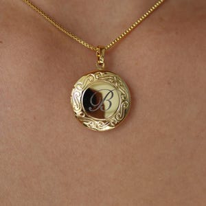 Peut inclure: Un collier avec un médaillon rond doré. Le médaillon porte la lettre "B" gravée et est orné d'un motif tourbillonnant. Il est suspendu à une fine chaîne dorée.