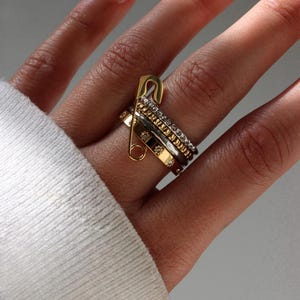 Puede incluir: Una mano muestra una colección de anillos dorados y plateados, incluyendo uno con diseño de imperdible. Los anillos presentan diversas texturas y adornos, como pequeños diamantes y patrones geométricos. Las joyas se destacan sobre un fondo suave y neutro.