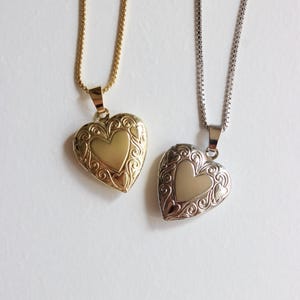 May include: Two heart-shaped lockets on chains. One is gold-toned, the other silver-toned. Both lockets have a heart shape in the center and ornate scrollwork around the edges. The chains are delicate and hang against a white background.