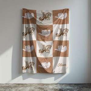May include: A brown and white checkered blanket featuring cowboy hats and riders on horseback. The blanket has a vintage, distressed look. The name "Oliver" is printed in cursive on the bottom right corner.