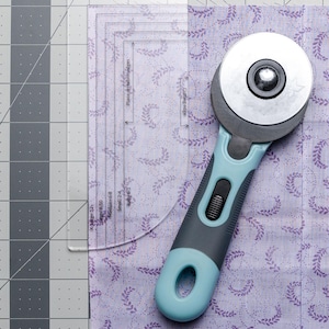 May include: A rotary cutter with a blue and gray handle rests on a piece of lavender fabric with a floral pattern. A clear quilting ruler is positioned over the fabric, and the cutting mat has a gray grid.