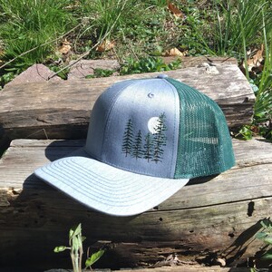 May include: A gray and green trucker hat with an embroidered forest and moon design. The front panel is gray with a forest and moon design, and the back is green mesh. The brim is gray.