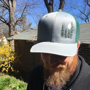 May include: A gray trucker hat with a green mesh back. The front panel features an embroidered design of pine trees and a full moon. The hat has a curved brim and a snapback closure.