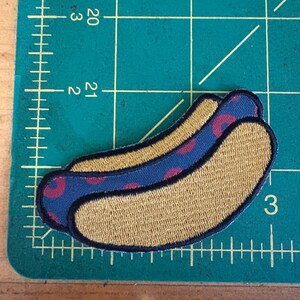 May include: Embroidered patch in the shape of a hot dog. The bun is tan, and the hot dog is blue with red polka dots. The patch has a black border and is on a green cutting mat.