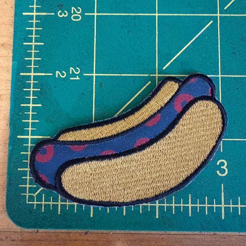 May include: Embroidered patch in the shape of a hot dog. The bun is tan, and the hot dog is blue with red polka dots. The patch has a black border and is on a green cutting mat.