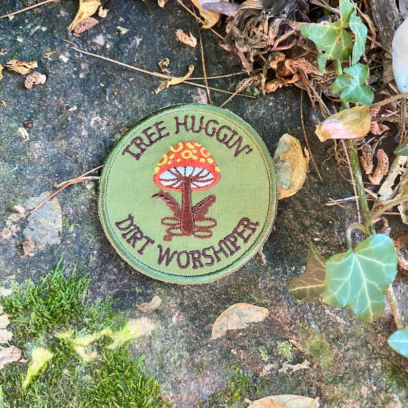 Pu&ograve; includere: Una toppa rotonda verde oliva con le parole "TREE HUGGIN'" e "DIRT WORSHIPER" ricamate in marrone. Un design di fungo colorato al centro, con un cappello rosso e arancione e uno stelo marrone.