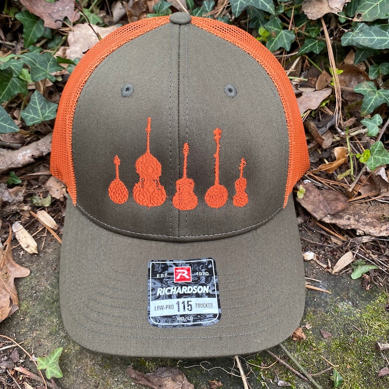 Pode incluir: Bon&eacute; de caminhoneiro verde-oliva e laranja com um design de instrumentos musicais. O bon&eacute; tem uma aba curva, uma parte traseira de malha e uma etiqueta "RICHARDSON 115 TRUCKER". A frente exibe instrumentos de cordas bordados em laranja.