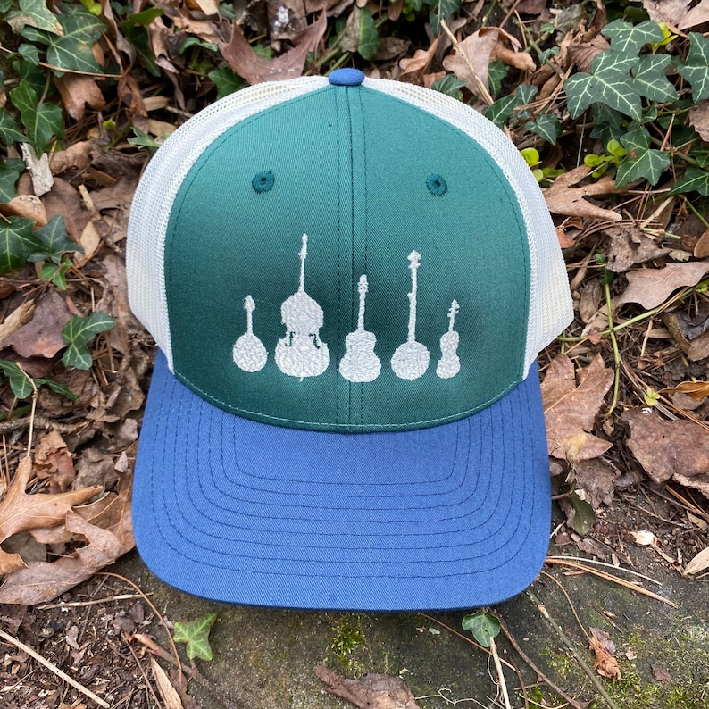 Pode incluir: Bon&eacute; de caminhoneiro verde e azul com costas em malha branca. O painel frontal apresenta um design branco de instrumentos musicais, incluindo um banjo, contrabaixo e guitarras. Estilo cl&aacute;ssico.