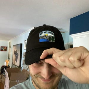 May include: Black baseball cap with a rectangular patch displaying the text "Aunt Life Grand" above a landscape scene with a lake, mountains, and trees. The cap is made of a dark fabric and has a curved brim.