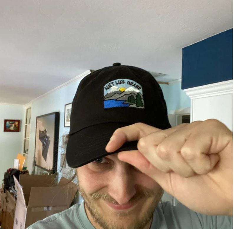 May include: Black baseball cap with a rectangular patch displaying the text "Aunt Life Grand" above a landscape scene with a lake, mountains, and trees. The cap is made of a dark fabric and has a curved brim.