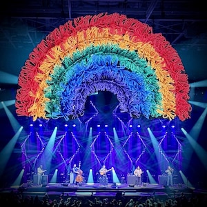 May include: A stage set with a large, colorful rainbow made of yarn. Below the rainbow, a band performs with guitars, a bass, and a violin. The stage is lit with blue and white spotlights, and the audience is visible in the foreground.