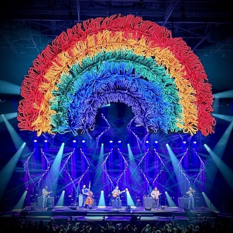 May include: A stage set with a large, colorful rainbow made of yarn. Below the rainbow, a band performs with guitars, a bass, and a violin. The stage is lit with blue and white spotlights, and the audience is visible in the foreground.