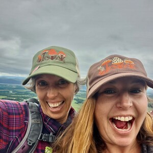 Pode incluir: Duas pessoas sorriem para a c&acirc;mera, usando bon&eacute;s de caminhoneiro com tema de cogumelos. Um bon&eacute; &eacute; verde-oliva com cogumelos laranja, o outro &eacute; marrom com cogumelos com bolinhas brancas. O fundo &eacute; um c&eacute;u nublado e uma paisagem distante.