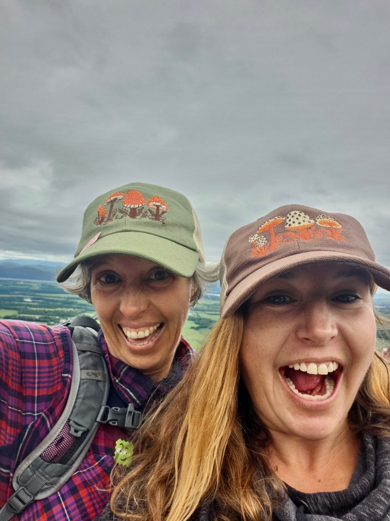 Pode incluir: Duas pessoas sorriem para a c&acirc;mera, usando bon&eacute;s de caminhoneiro com tema de cogumelos. Um bon&eacute; &eacute; verde-oliva com cogumelos laranja, o outro &eacute; marrom com cogumelos com bolinhas brancas. O fundo &eacute; um c&eacute;u nublado e uma paisagem distante.