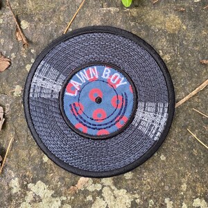 May include: A circular patch resembling a vinyl record. The outer edge is black, with a textured gray surface. The center features a blue circle with the words "LAWN BOY" and red donut-shaped designs. The patch is likely an embroidered or fabric item.