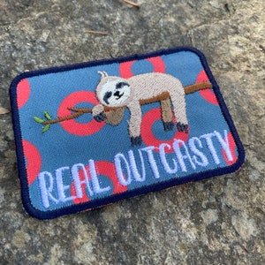 May include: Embroidered patch with a sloth design. The patch has a blue background with red circles and the words "REAL OUTCASTY" in white. The sloth is light brown and the branch is brown. The patch is rectangular.