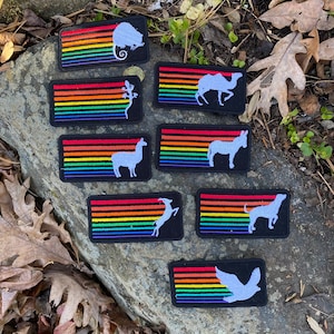 May include: Rectangular black patches with a rainbow striped design and various animal silhouettes in white. Animals include a mouse, lizard, camel, donkey, llama, deer, dog, and dove. Patches are displayed on a rock surface.