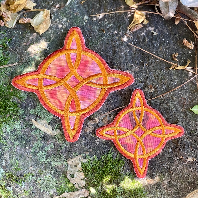 May include: Two embroidered patches with a Celtic knot design. The patches feature a gold-colored knot over a pink and orange background, with a red border. One patch is larger than the other.