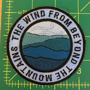 May include: Circular embroidered patch with a mountain landscape design in blue and green hues. The text "THE WIND FROM BEYOND THE MOUNTAINS" is embroidered in white around the edge. The patch is on a green surface.