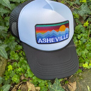 Peut inclure: Casquette de camionneur avec une visi&egrave;re et un dos en filet noirs, un panneau avant bleu clair et un &eacute;cusson repr&eacute;sentant une montagne color&eacute;e et un coucher de soleil avec le mot "ASHEVILLE" en lettres blanches. D&eacute;tail de corde noire.